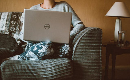 A women wearings leggings and a sweater sits comfortably on couch with her laptop.